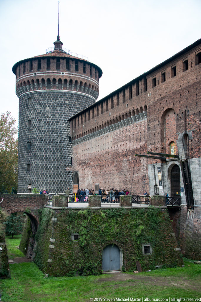 Round stone-faced corner tower beside a long brick wall, gateway, bridge, and grassy moat at Castello Sforzesco.