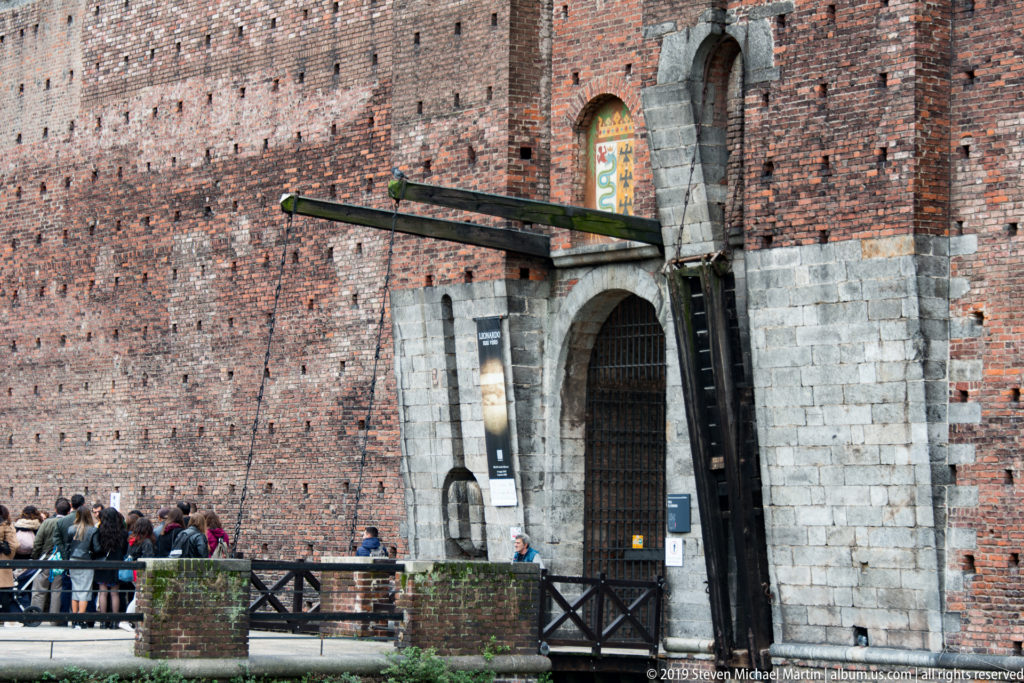 Arched castle gateway framed in pale stone within a massive brick wall, with a bridge and visitors at Castello Sforzesco.