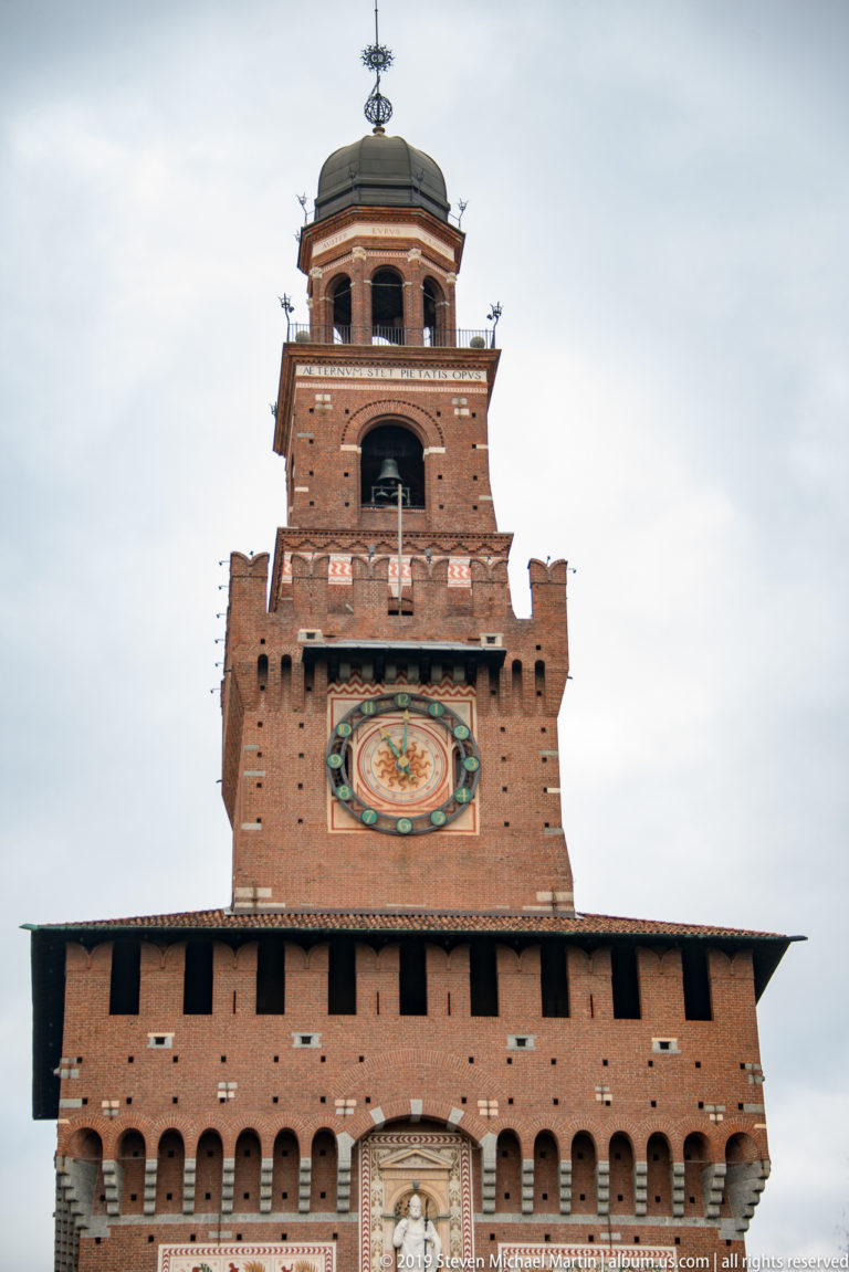 Upper Torre del Filarete with circular clock, brick belfry-like stage, domed cap, and projecting roofline against a cloudy sky.