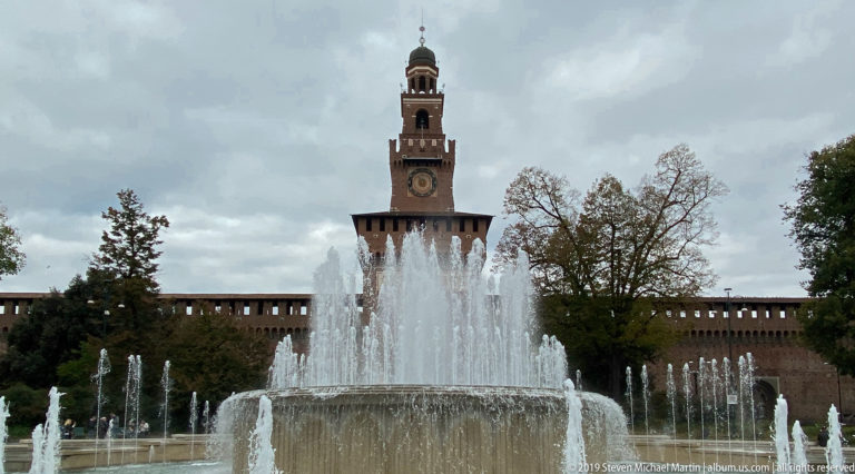 Fountain jets in front of the brick Torre del Filarete, with its clock and lantern, at Castello Sforzesco in Milan.