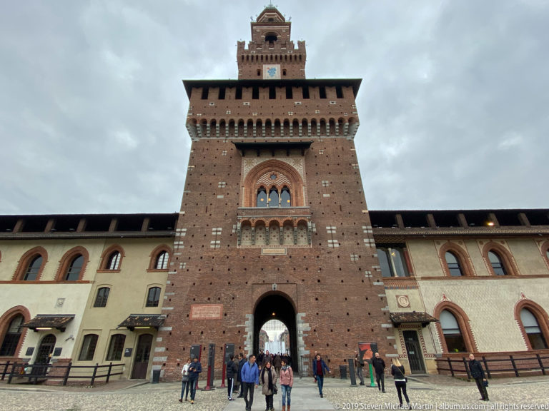 Brick Torre del Filarete with clock and large arched gateway, viewed from inside Castello Sforzesco with visitors below.