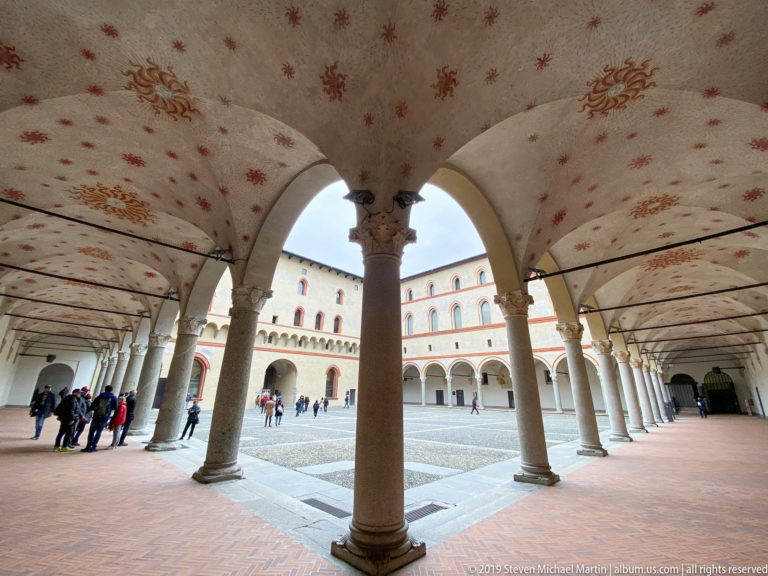 Stone columns and broad arches support painted vaults along an inner courtyard arcade at Castello Sforzesco.