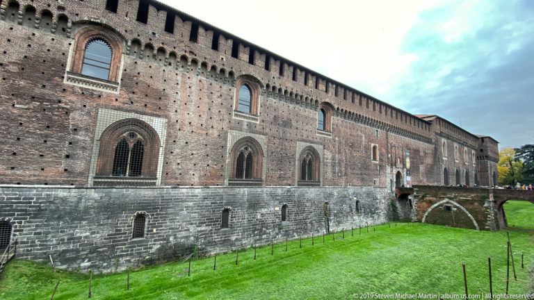 Long brick wall of Castello Sforzesco above a grassy moat, with pointed-arch windows and a dark stone base.