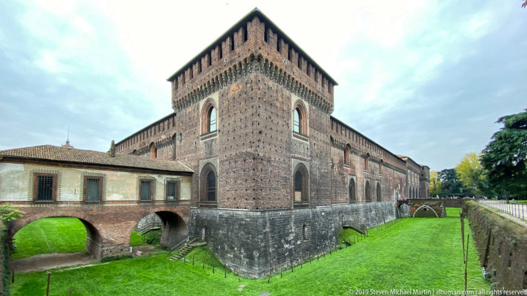 Square brick corner tower rising above the grassy moat, with arched bridges beside Castello Sforzesco in Milan.