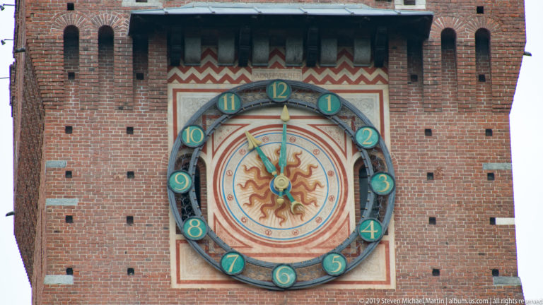 Large circular clock with green numbered medallions and a radiant sun face set in a painted panel on the Torre del Filarete.