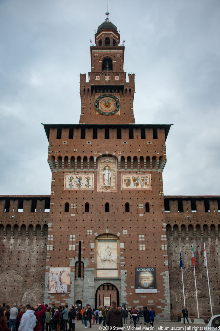Frontal view of the Torre del Filarete above Castello Sforzesco’s main gateway, with clock, heraldry, statue, and visitors.