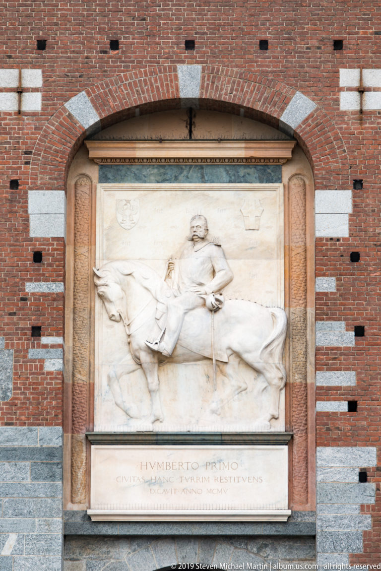 Marble relief of a mounted ruler above an inscription naming Umberto Primo, set into a red-brick castle wall.