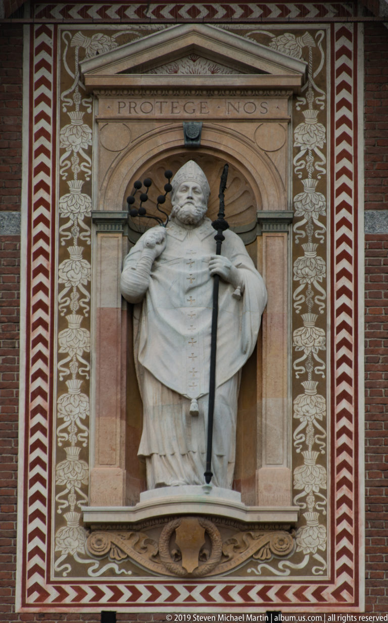 White stone bishop holding a crozier in an arched niche beneath the inscription PROTEGE NOS on the Torre del Filarete.