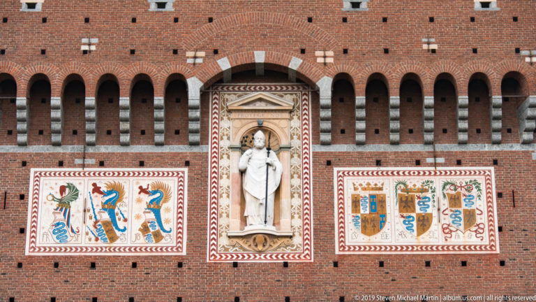 White robed figure in a tall niche between colorful heraldic panels with serpents and eagles on the Torre del Filarete.