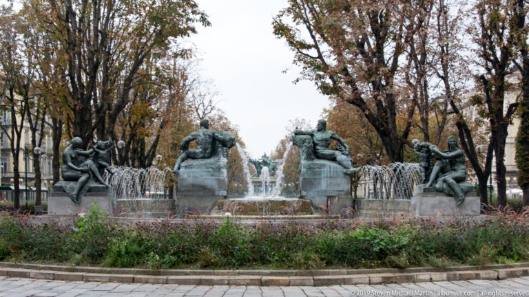 Wide view of a large sculptural fountain with bronze figures, multiple water jets, trees, and landscaped planting around the basin.