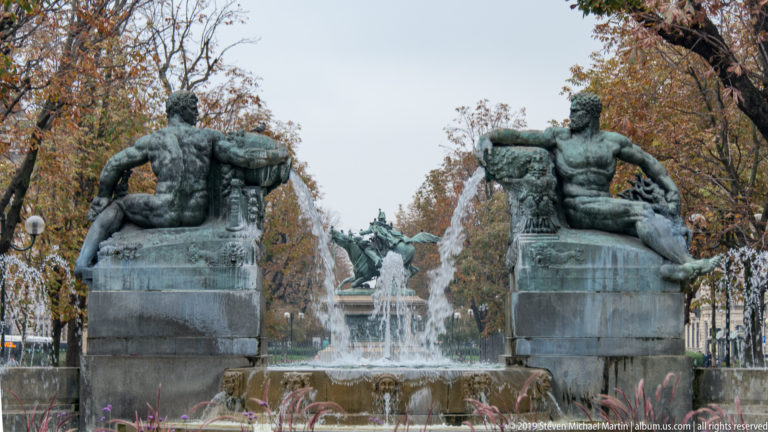 Rear view of two monumental bronze male figures on fountain plinths framing a central basin and distant equestrian monument.