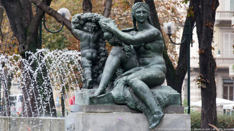 Bronze fountain group of a seated woman and child on a stone base, with arcing water jets behind them.