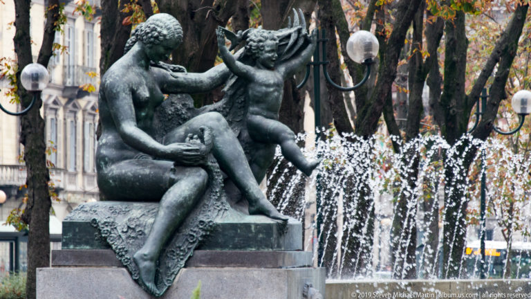 Bronze fountain sculpture of a seated woman with a child, backed by white water jets and autumn trees.