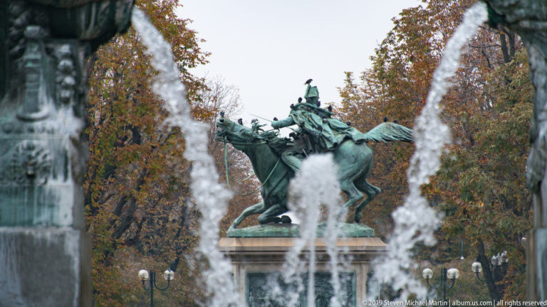Bronze equestrian statue on a pedestal seen between tall fountain jets, with autumn trees in the background.