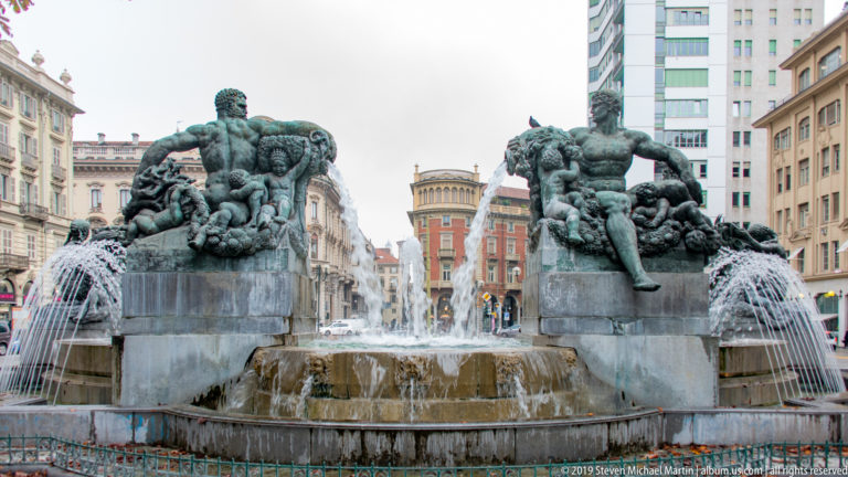 Monumental city fountain with two large bronze male figures on stone plinths and multiple water jets around a central basin.