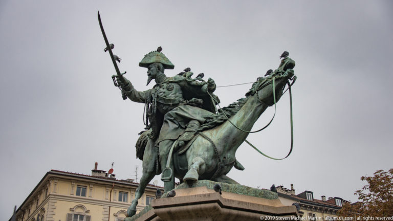 Green bronze equestrian monument of a uniformed rider with raised arm and plumed headgear against a gray sky.