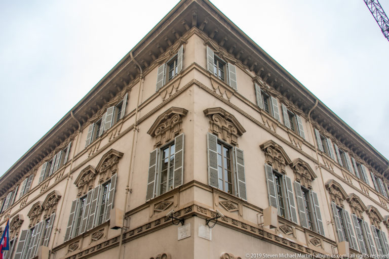 Low-angle corner view of a pale historic palazzo with shuttered windows, carved pediments, and a heavy projecting cornice.