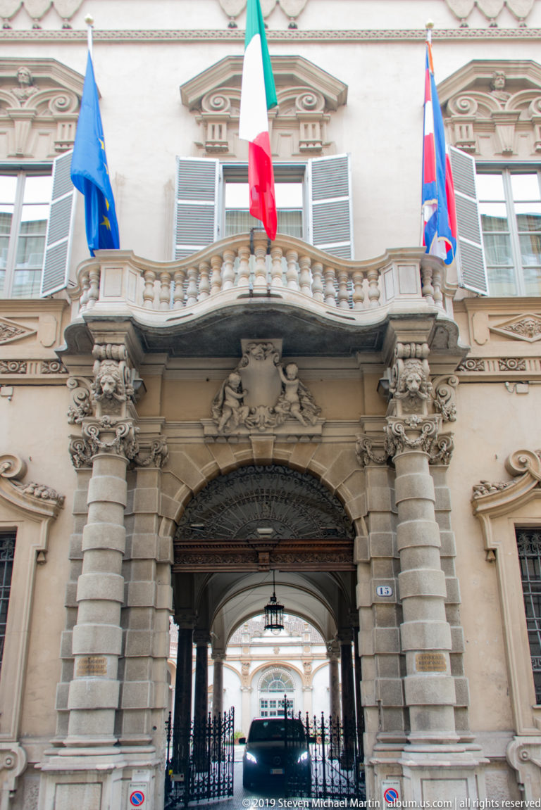 Large arched palace entrance with rusticated stonework, sculpted balcony, official flags, and a passage leading to an inner court.