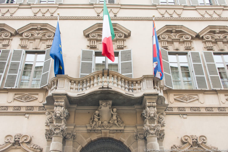 Ornate stone balcony with Italian, European Union, and Piedmont flags above carved portal decoration and shuttered windows.