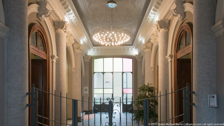 Ornate Turin entrance hall with chandelier, classical columns, paired wooden doors, wrought-iron gate, and bright rear window.