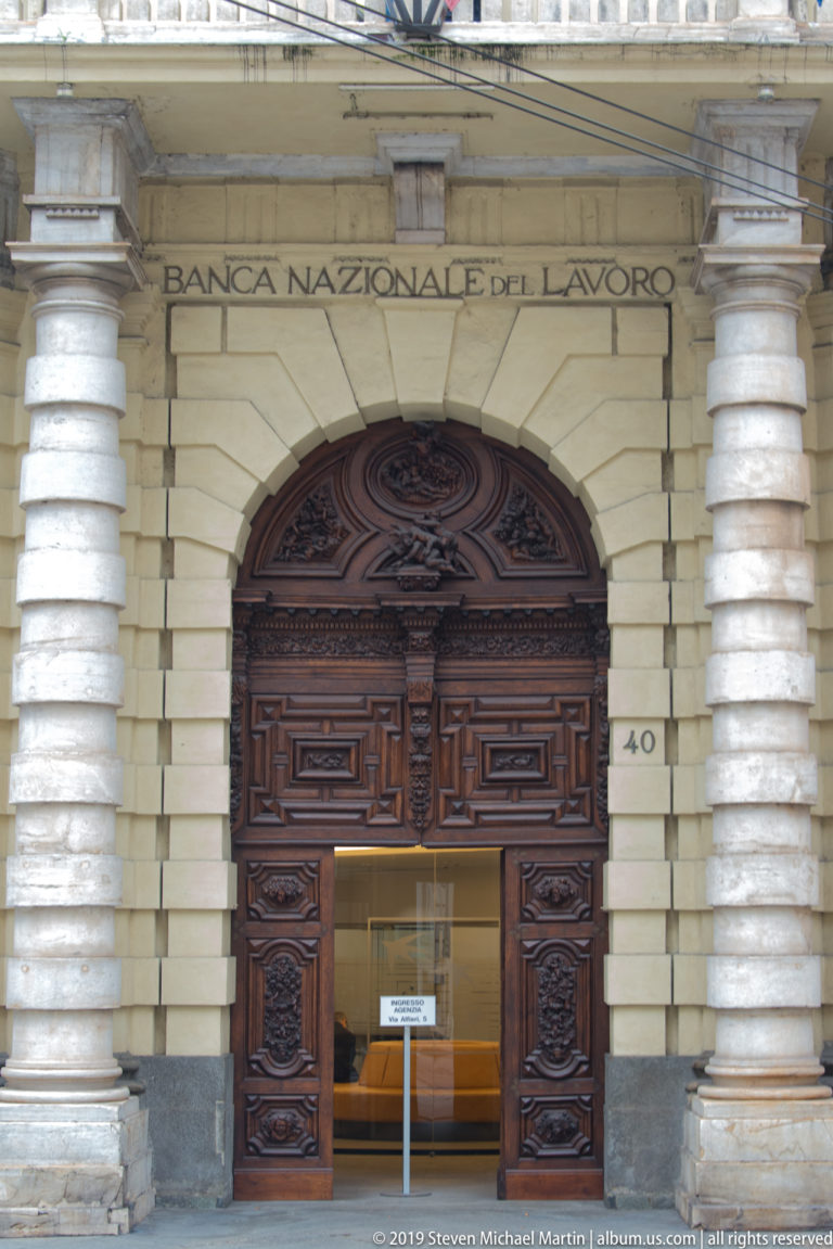 Banca Nazionale del Lavoro entrance with carved name, rusticated stonework, columns, and a large arched wooden doorway.