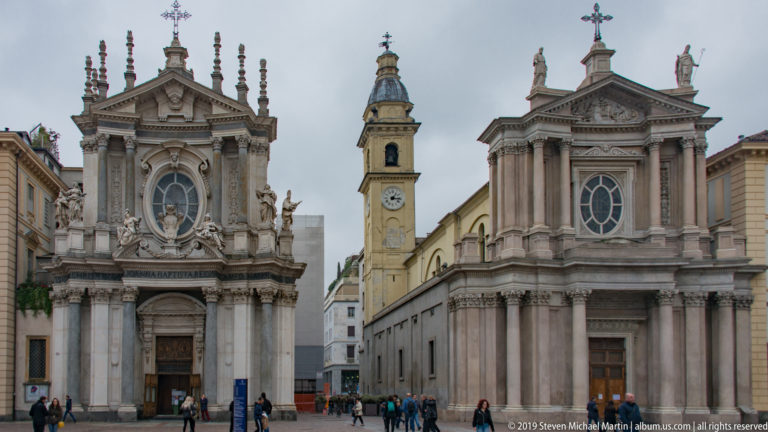 Close frontal view of Piazza San Carlo’s twin churches with a clock tower visible in the narrow gap between them.