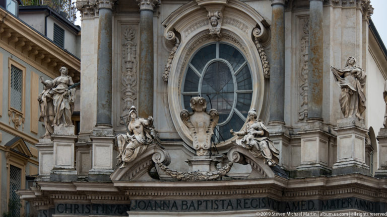 Baroque church façade detail with an oval window, columns, large stone figures, scrollwork, and a Latin inscription below.