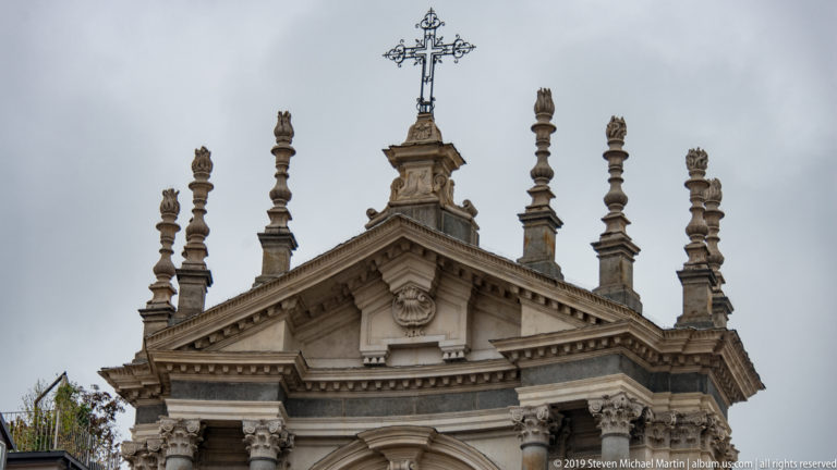 Upper church façade with triangular pediment, sculpted figures, pinnacles, and a metal cross against a gray sky.