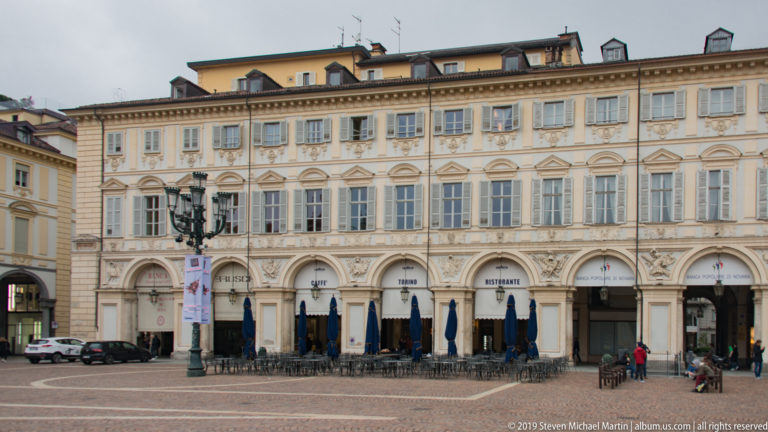 Pale historic façade with shuttered windows, ground-floor arches, café awnings, and outdoor seating on Piazza San Carlo.