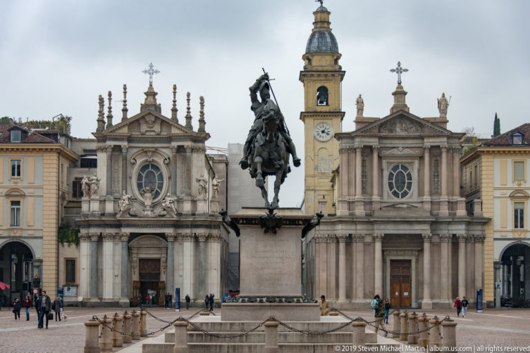 Twin church façades of Piazza San Carlo behind the bronze equestrian monument, with a clock tower rising between them.