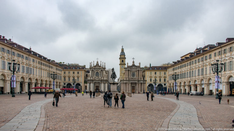 Wide cobbled Piazza San Carlo with arcaded palaces, twin churches, and a central equestrian monument in the distance.