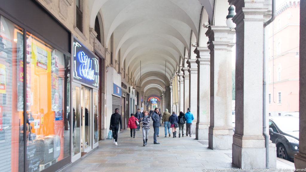 Pedestrians walking beneath a vaulted Turin shopping arcade with storefronts on the left and stone piers along the street.