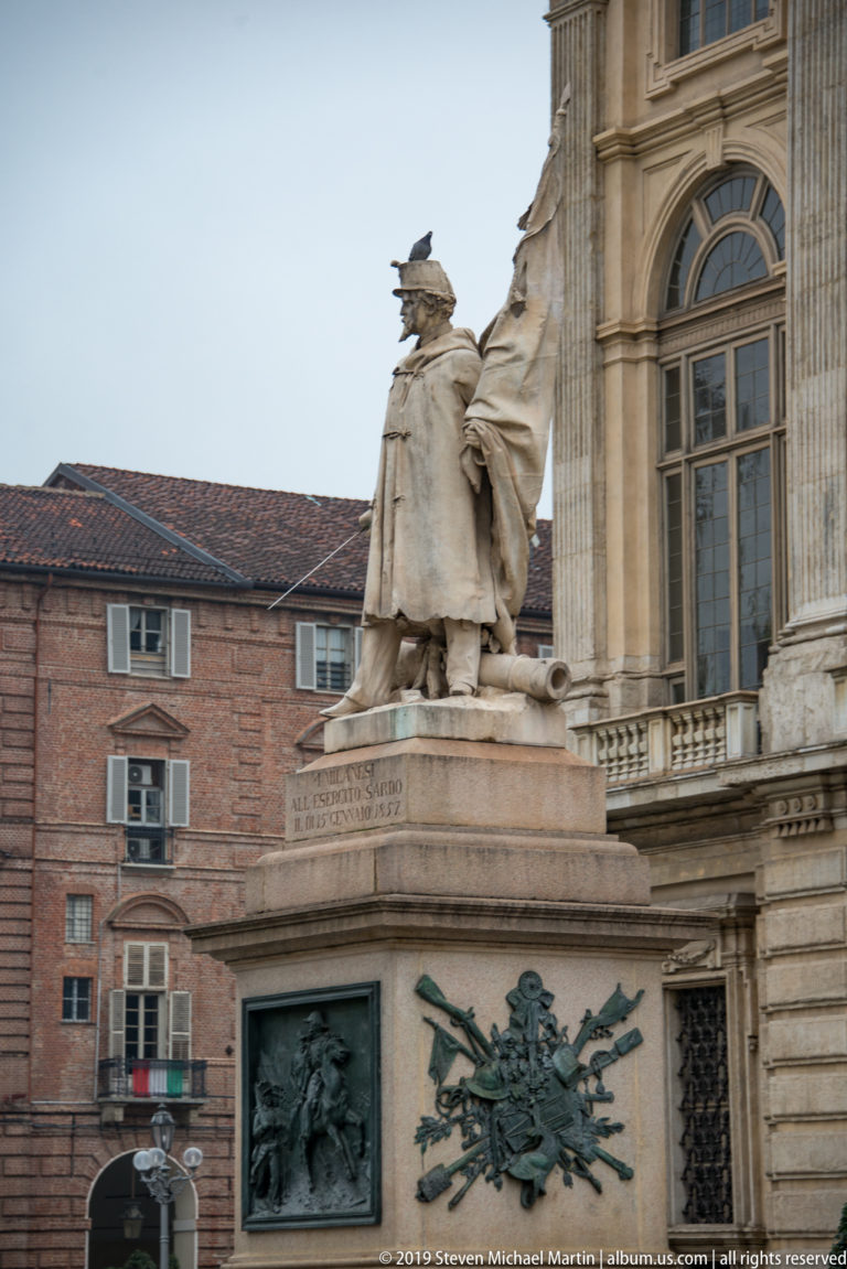 Seated stone statue on a high pedestal above a rectangular bronze relief beside a palace façade.