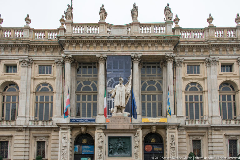 Close view of an ornate palace entrance with columns, arched windows, a standing statue, flags, and relief panels.