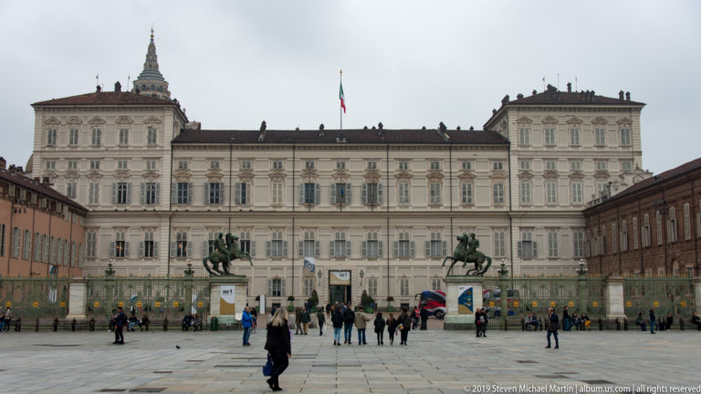 Palazzo Reale façade across Piazza Castello with the Italian flag centered above the entrance and people in the foreground.