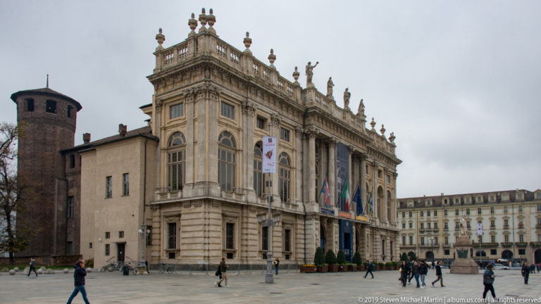 Ornate Baroque palace façade with tall windows, rooftop statues, flags, and pedestrians crossing the square in front.