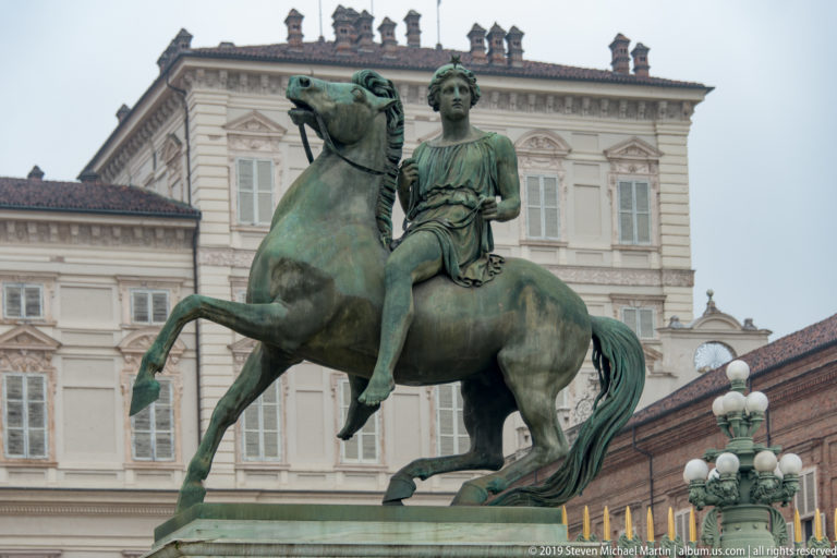 Green-patinated bronze rider on a horse in front of a pale historic palace façade.