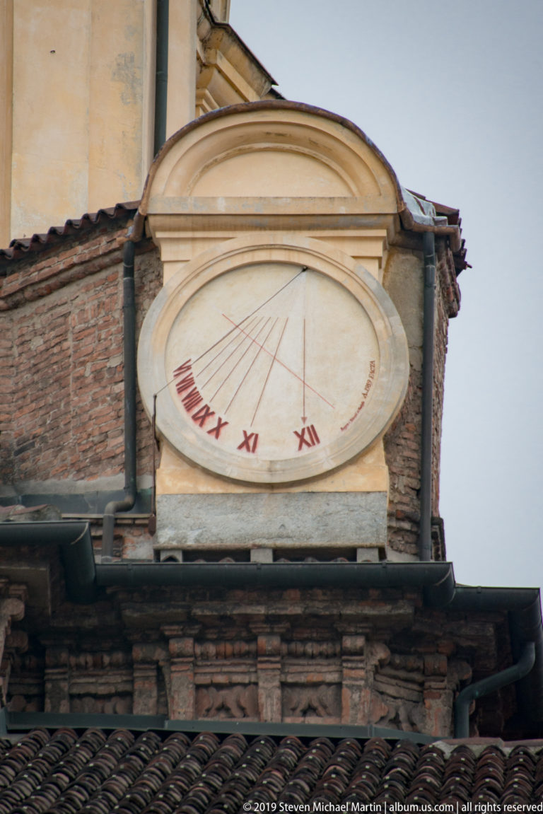 Wall-mounted sundial with radiating hour lines and red Roman numerals above a dark tiled roof.