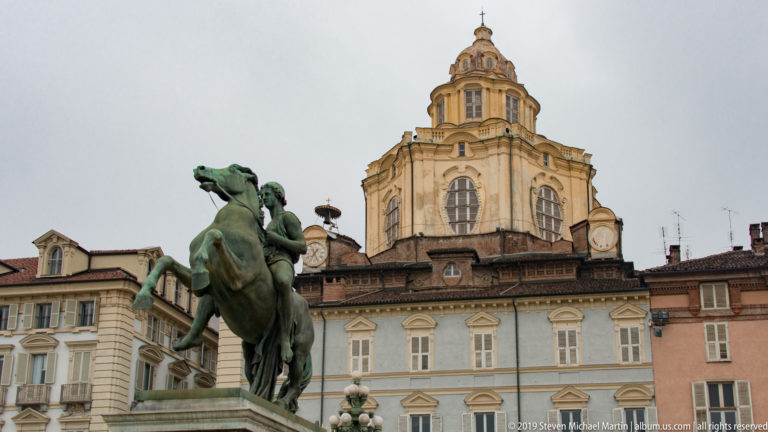 Bronze equestrian statue seen from below with an ornate church dome and historic façades rising behind it.