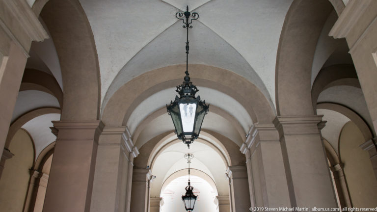 Vaulted covered portico with repeated arches and black hanging lanterns receding toward a bright opening.