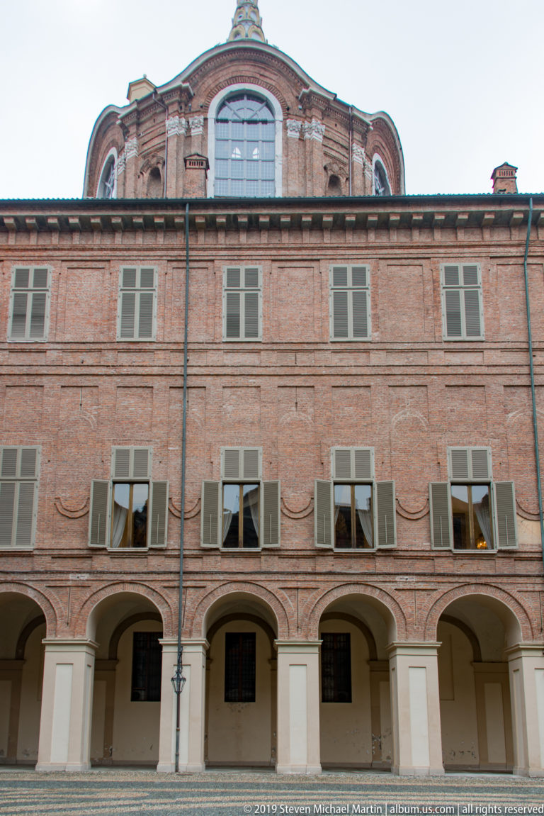 Brick palace wing with repeated ground-floor arches, shuttered windows, and a raised central roof pavilion.