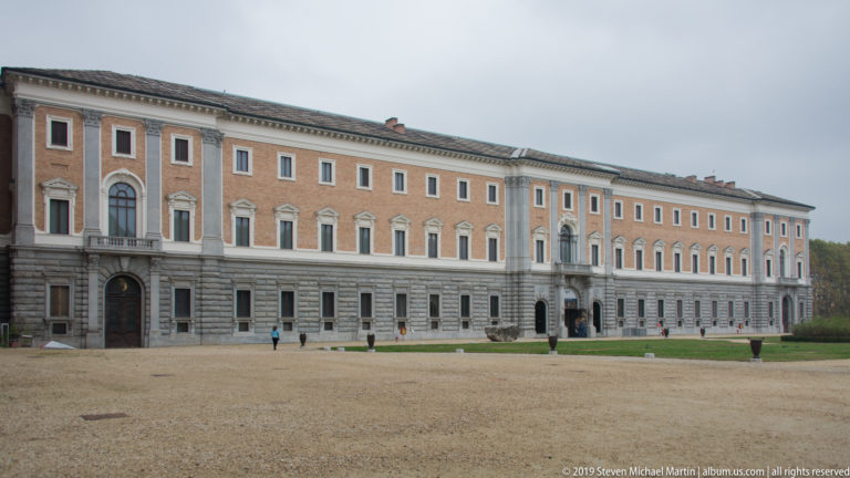 Long palace façade with rusticated ground floor and rows of windows facing a broad gravel courtyard under an overcast sky.