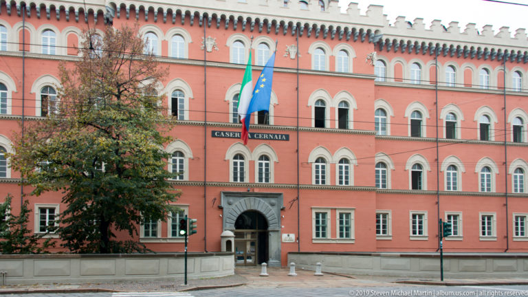 Red Caserma Cernaia façade with arched windows, crenellated roofline, central entrance, and Italian and EU flags.