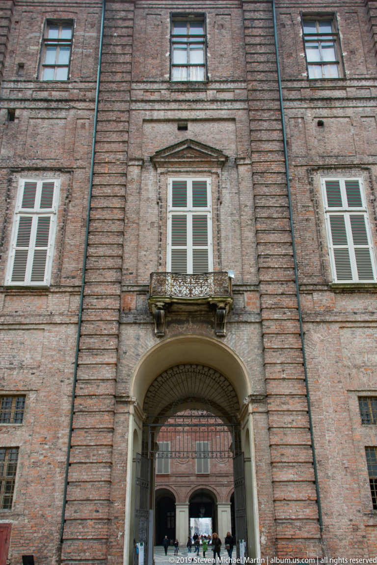 Brick palace façade with shuttered windows, a small stone balcony, and a large arched gateway opening into an inner courtyard.