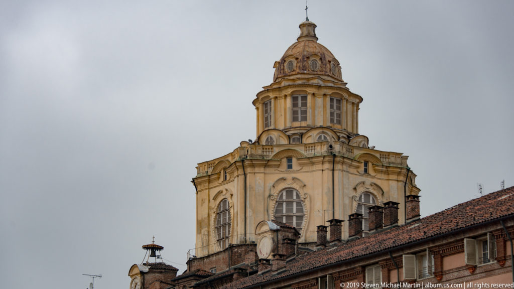 Ornate Baroque dome with arched windows and lantern rising above tiled rooftops under a gray sky.