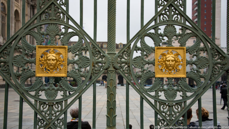 Two gilded Medusa-like masks repeated across ornate green wrought-iron gate panels in Turin.