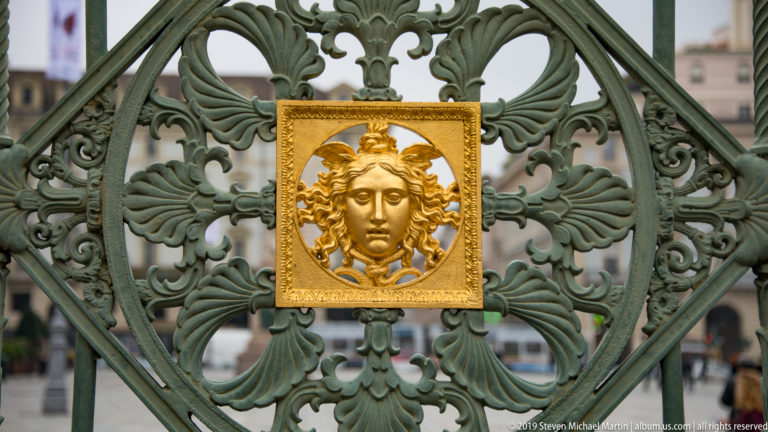 Gilded Medusa-like head in a square panel centered within ornate green-painted wrought-iron gatework.