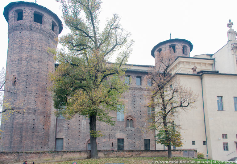 Two cylindrical brick towers of Palazzo Madama rising behind trees beside a pale adjoining building.