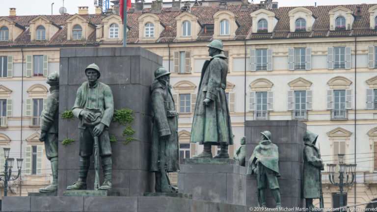 Life-size bronze figures in coats and hats arranged around a dark stone monument in a Turin square.
