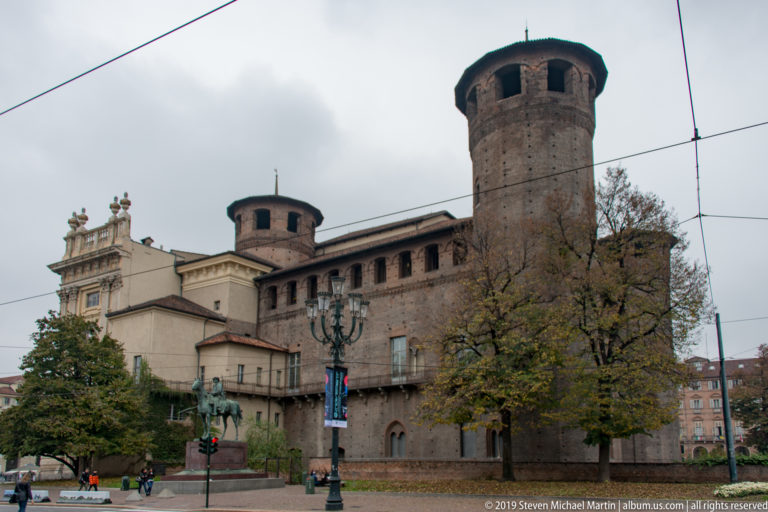 Wide view of Palazzo Madama’s medieval brick towers and adjoining buildings beneath tram wires in Piazza Castello.
